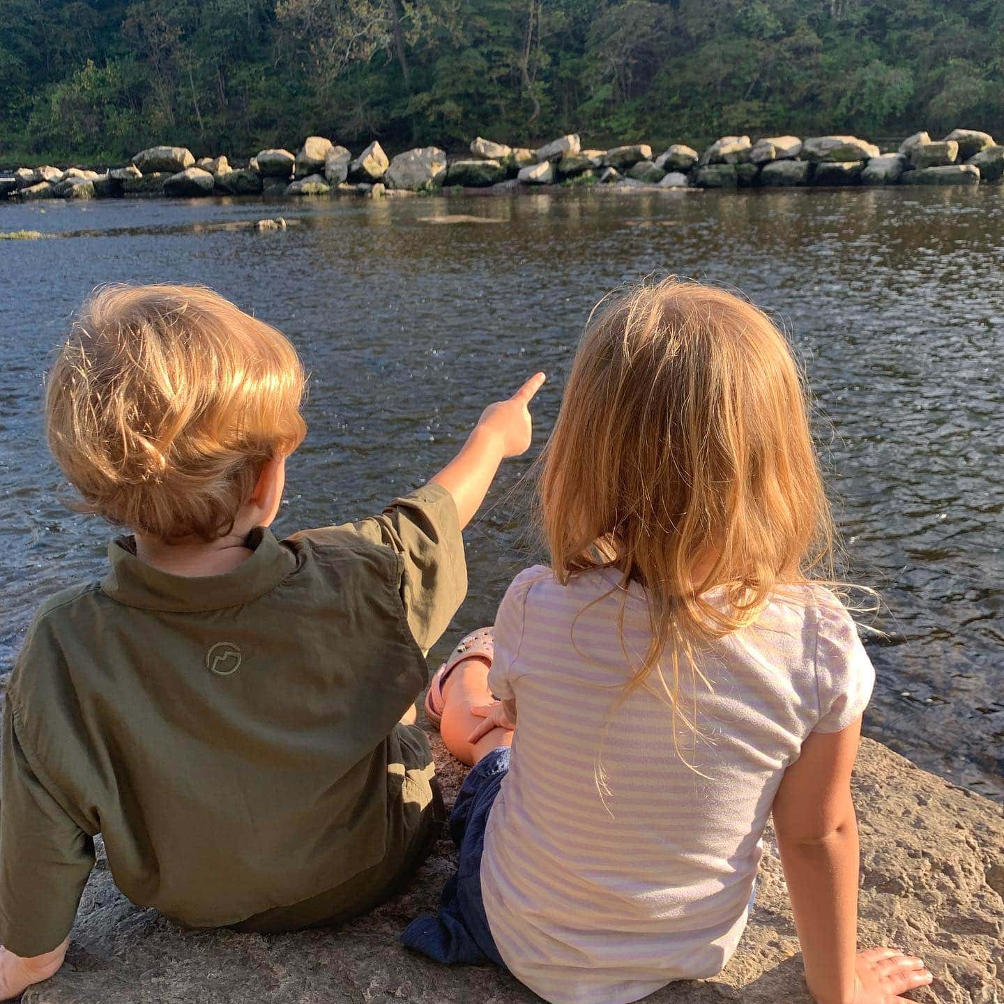 Simply Learning Together Lakeside View A young boy and girl sitting on rocks by a river, enjoying nature and outdoor adventure.